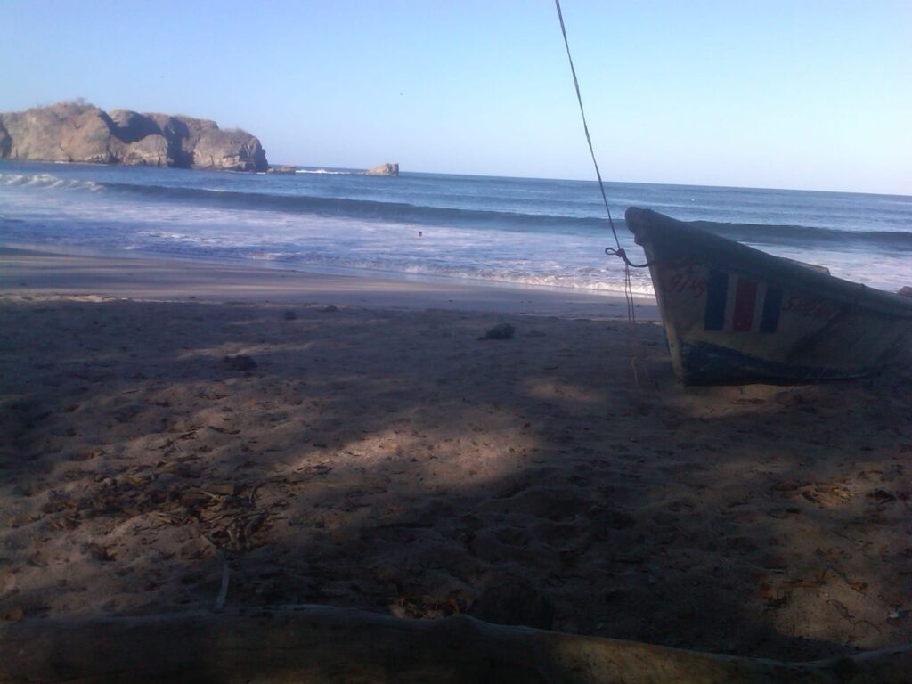 Fishing boat on a quiet Nosara beach with waves and a rocky point. Arrive easily with our Nosara shuttle service.